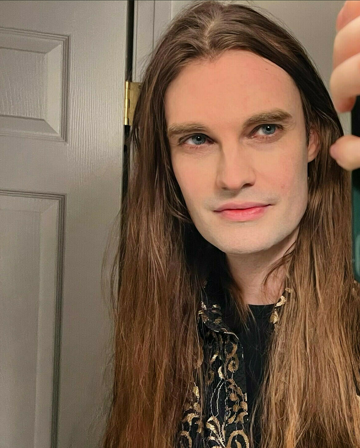 An androgynous white person with long brown hair and blue eyes takes a selfie.